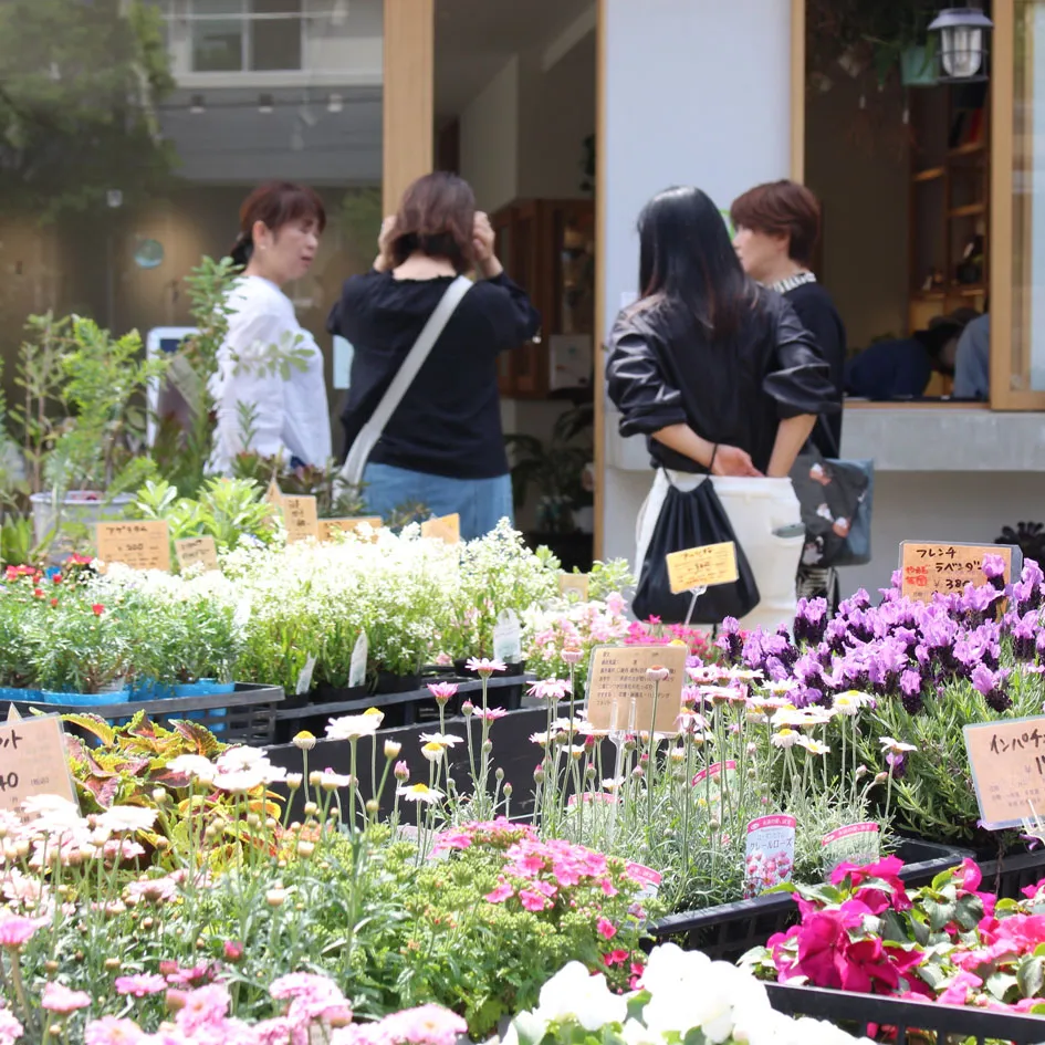 カラフルな花々が並ぶ花ノ停留所屋外店舗で、数人の客が植物を選んでいる様子。